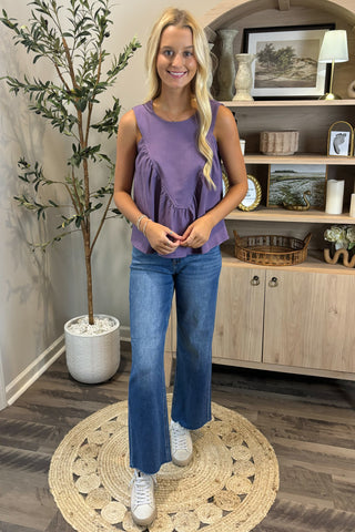 Woman in a purple top and blue jeans standing in a room with a plant and wooden cabinet.