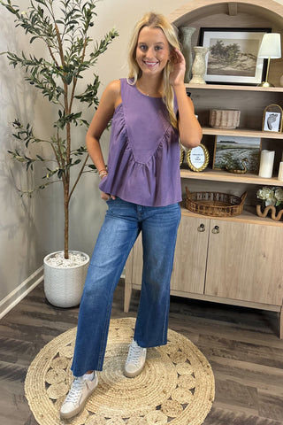 Woman wearing a purple top and blue jeans standing in a room with a plant and shelves.