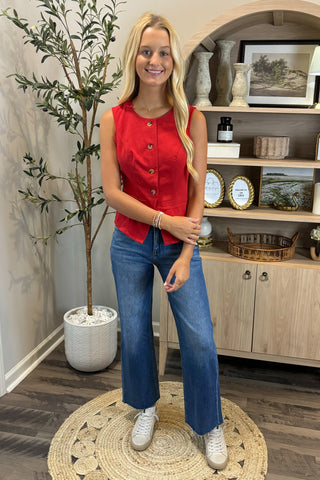 Woman in a red sleeveless top and blue jeans standing in a room with a plant and shelves.