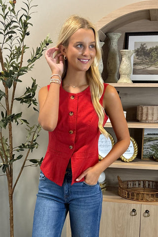 Woman wearing a red sleeveless top and blue jeans standing indoors with decorative elements in the background.