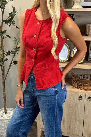 Person wearing a red sleeveless denim jacket and blue jeans indoors.