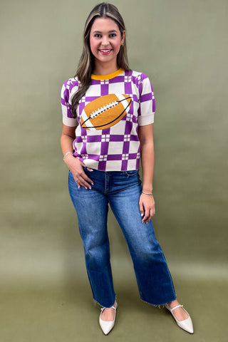 Woman wearing a purple checkered shirt with a football graphic and blue jeans on a beige background