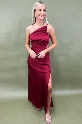 Woman wearing a burgundy one-shoulder dress with a slit on a plain background