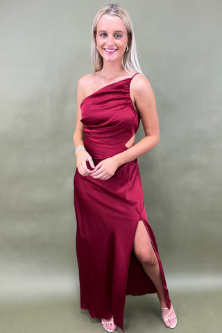 Woman wearing a burgundy one-shoulder dress with a slit on a plain background