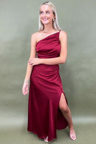 Woman wearing a burgundy one-shoulder dress with a slit on a plain background