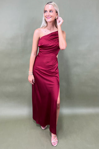 Woman wearing a burgundy one-shoulder dress with a slit on a plain background