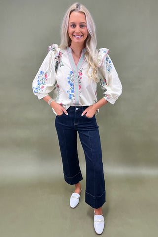 Woman wearing a white blouse with floral embroidery and dark jeans on a plain background