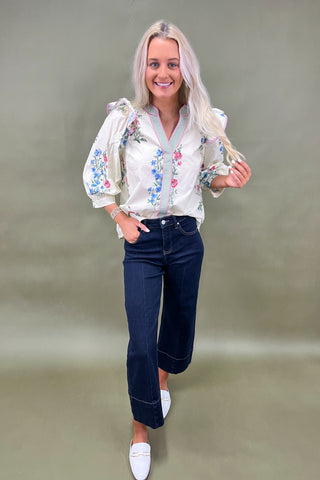 Woman wearing a white blouse with floral embroidery and dark jeans on a plain background
