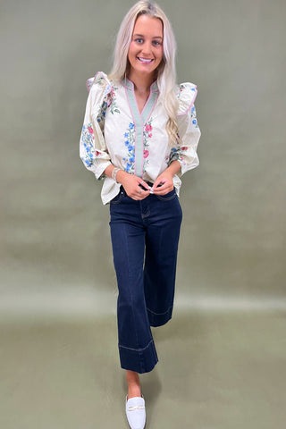 Woman wearing a white blouse with floral embroidery and dark jeans on a plain background