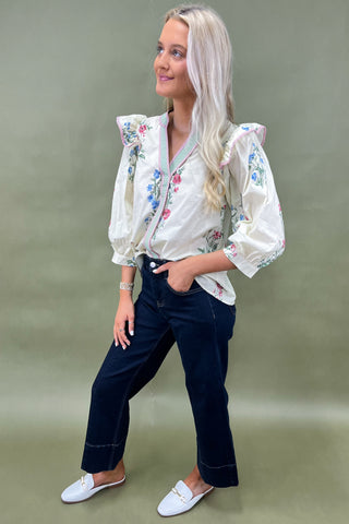 Woman wearing a white blouse with floral embroidery and dark pants against a green background