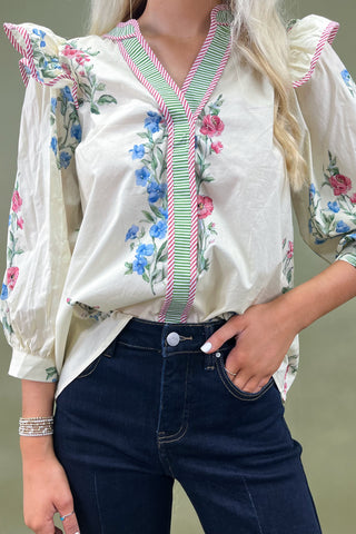 Woman wearing a white blouse with floral embroidery and blue jeans against a neutral background