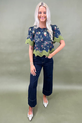 Woman wearing a floral blouse with green trim and dark jeans on a plain background
