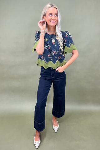 Woman wearing a navy floral top with green trim and dark jeans on a plain background