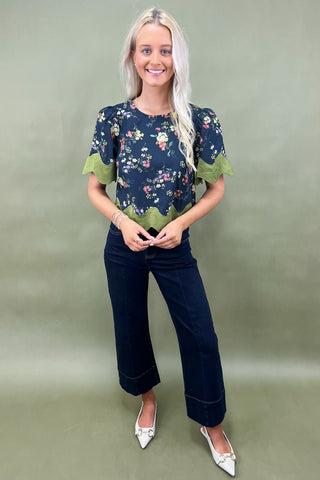 Woman wearing a floral blouse with green trim and dark jeans on a plain background