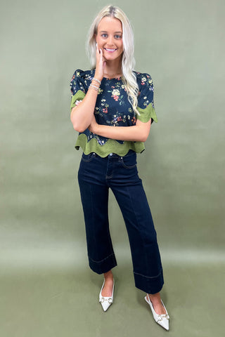 Woman wearing a floral blouse with green trim and dark jeans on a plain background