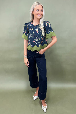Woman wearing a floral blouse with green trim and dark jeans on a plain background
