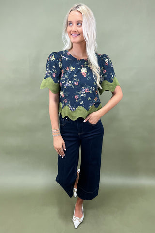 Woman wearing a floral blouse with green trim and dark jeans on a plain background