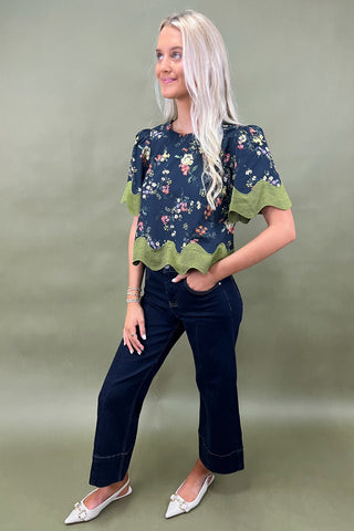 Woman wearing a navy floral top with green trim and dark jeans on a plain background