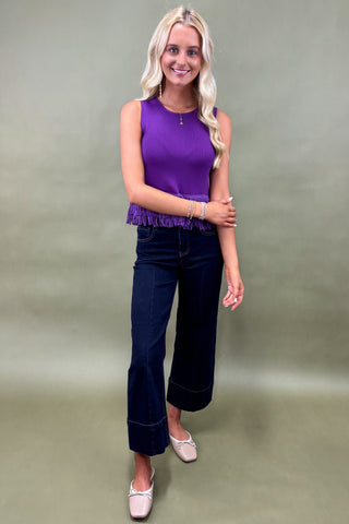 Woman wearing a purple sleeveless top and dark blue jeans against a plain background