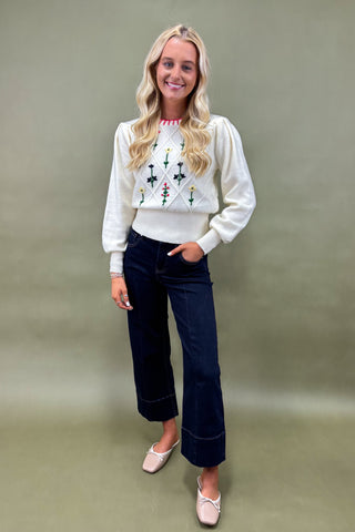 Woman wearing a white sweater with a pattern and dark jeans against a plain background