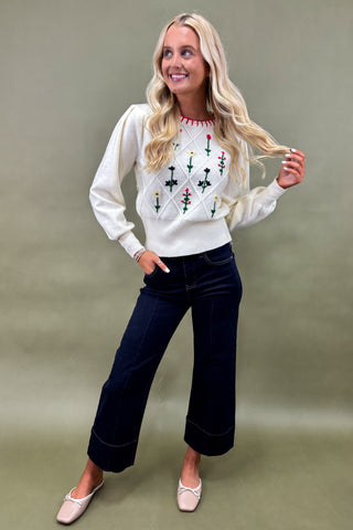 Woman wearing a white sweater with red and green patterns and dark blue jeans on a plain background