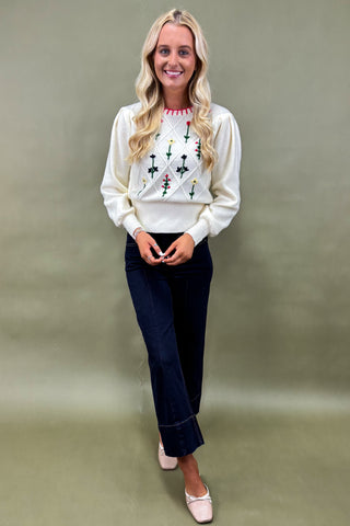 Woman wearing a white sweater with floral patterns and dark jeans against a plain background
