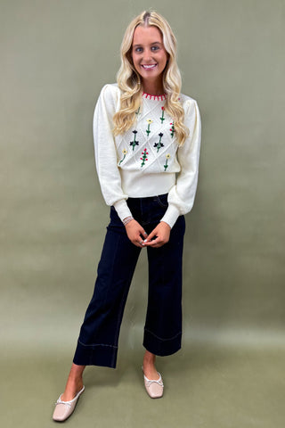 Woman wearing a white sweater with floral patterns and dark jeans on a plain background