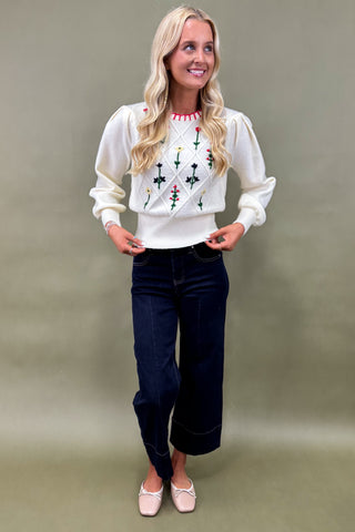 Woman wearing a white embroidered blouse and dark jeans against a plain background