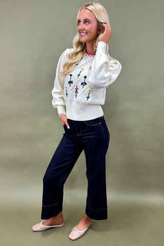 Woman wearing a white sweater with floral patterns and dark jeans on a plain background