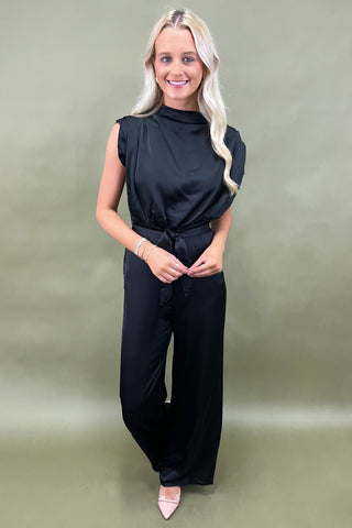 Woman wearing a black sleeveless top and wide-leg pants against a green background