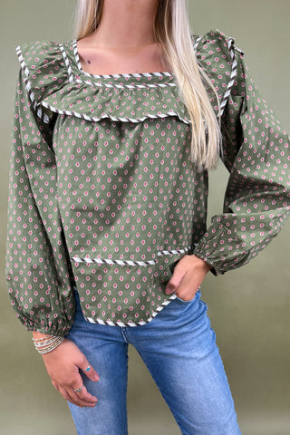 Woman wearing a green patterned blouse with ruffled details and blue jeans on a plain background