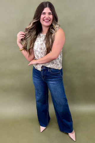 Woman wearing a sleeveless cream floral top with brown print, paired with dark wash wide leg jeans and nude pointed heels, boutique fashion photography on olive backdrop