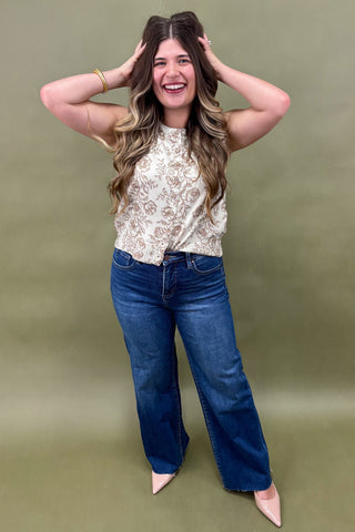 Woman wearing a sleeveless cream floral top with brown print, paired with dark wash wide leg jeans and nude pointed heels, boutique fashion photography on olive backdrop