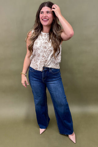 Woman wearing a sleeveless cream floral top with brown print, paired with dark wash wide leg jeans and nude pointed heels, boutique fashion photography on olive backdrop