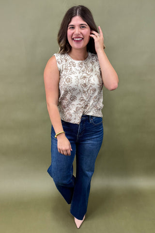 Woman wearing a sleeveless cream floral top with brown print, paired with dark wash wide leg jeans and nude pointed heels, boutique fashion photography on olive backdrop
