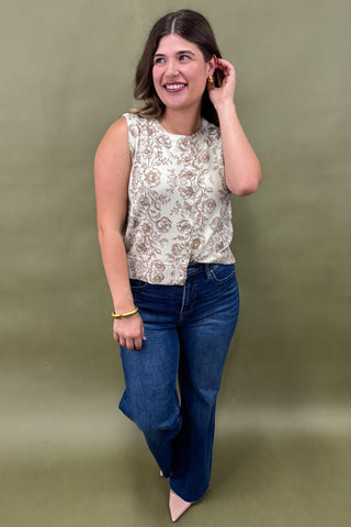 Woman wearing a sleeveless cream floral top with brown print, paired with dark wash wide leg jeans and nude pointed heels, boutique fashion photography on olive backdrop