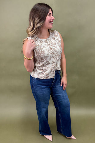Woman wearing a sleeveless cream floral top with brown print, paired with dark wash wide leg jeans and nude pointed heels, boutique fashion photography on olive backdrop
