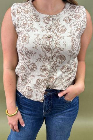 Woman wearing a sleeveless cream floral top with brown print, paired with dark wash wide leg jeans and nude pointed heels, boutique fashion photography on olive backdrop