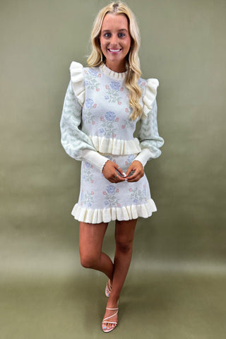 Woman wearing a pastel floral knit sweater dress with ruffle trim and puff sleeves, styled with strappy nude heels, boutique fashion photography on olive green backdrop