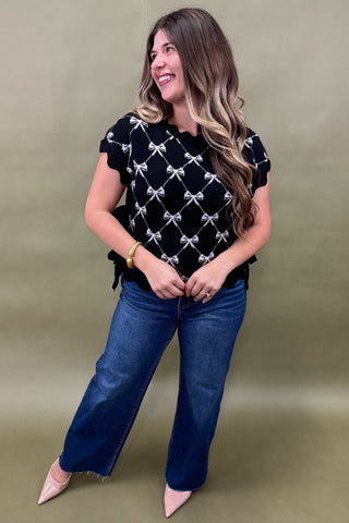 Woman wearing a black top with white patterns and blue jeans against a beige background