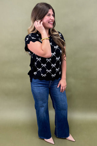 Woman wearing a black top with white patterns and blue jeans against a green background