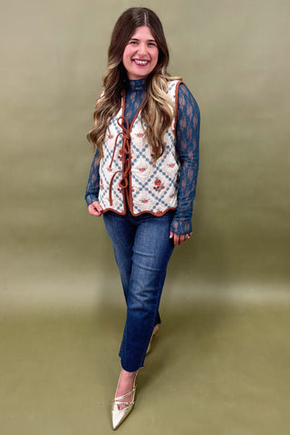 Woman wearing a patterned vest over a blue shirt with jeans on a green background