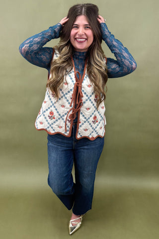 Woman wearing a patterned vest over a blue top and jeans on a green background