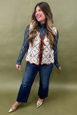 Woman wearing a patterned vest over a lace top with blue jeans against a green background