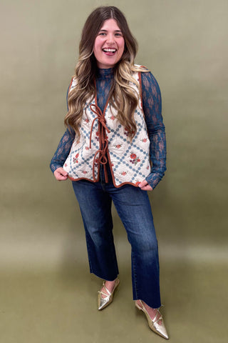 Woman wearing a patterned vest over a lace top with jeans and gold shoes on an olive green background