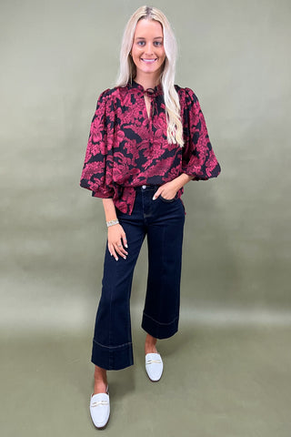 Woman wearing a red floral blouse and black pants against a plain background