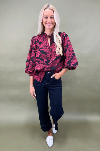 Woman wearing a red floral blouse and dark jeans against a plain background