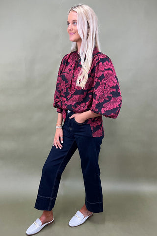 Woman wearing a red and black patterned blouse with navy pants against a plain background