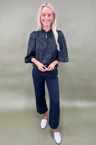 Woman wearing a dark blouse and jeans with white shoes on a plain background