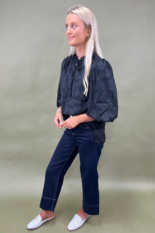 Woman wearing a dark patterned blouse and navy pants against a plain background